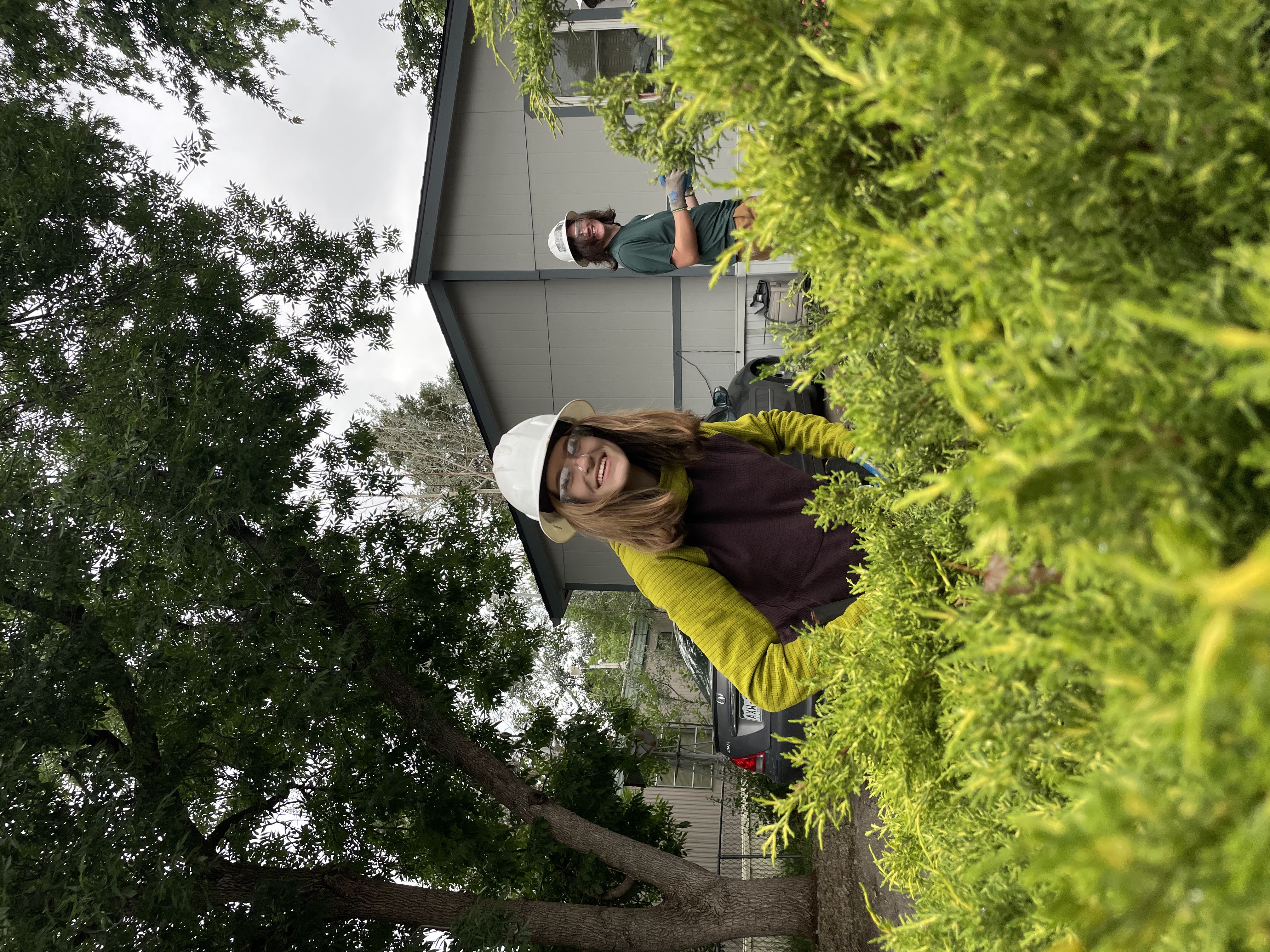 A crew member doing fire mitigation work at a home. She is smiling and surrounded by bushes. 