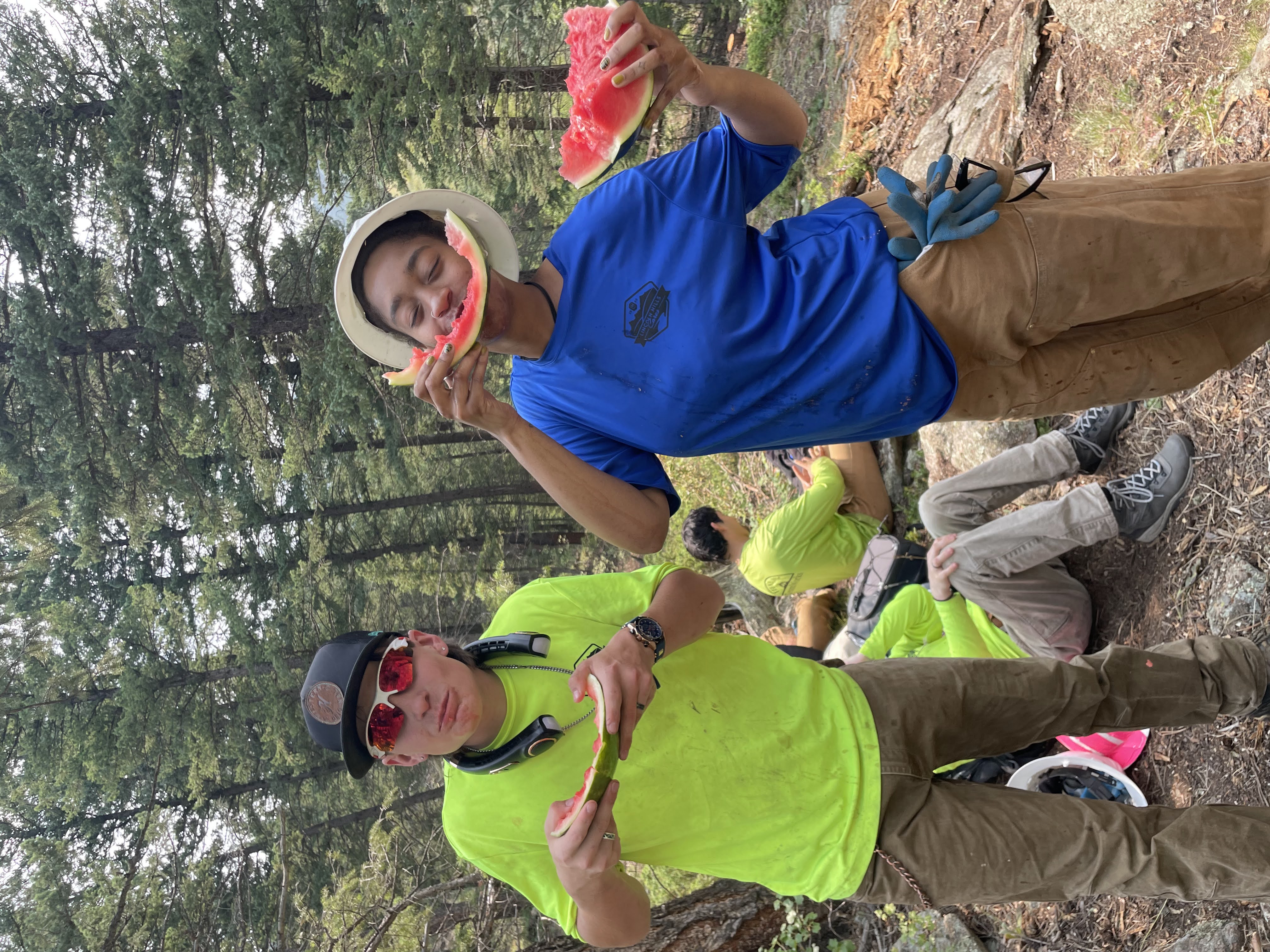 Two crew members laughing and eating watermelon. One crew member is holding the rind in front of her mouth to look like a smile. 