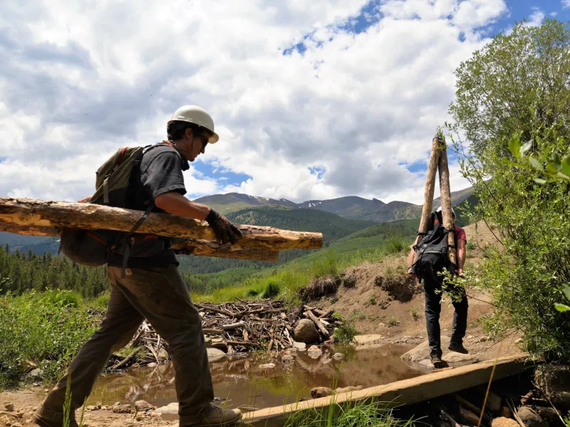 young people in hardhats carry cut logs over a mountain trail in Colorado