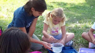 An environmental educator helps a student explore bugs while sitting in the grass under a tree