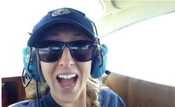 A young woman smiles with excitement, dressed in a navy Colorado Parks and Wildlife uniform, sunglasses, and large blue headphones. She is in the back seat of a small fixed wing airplane.