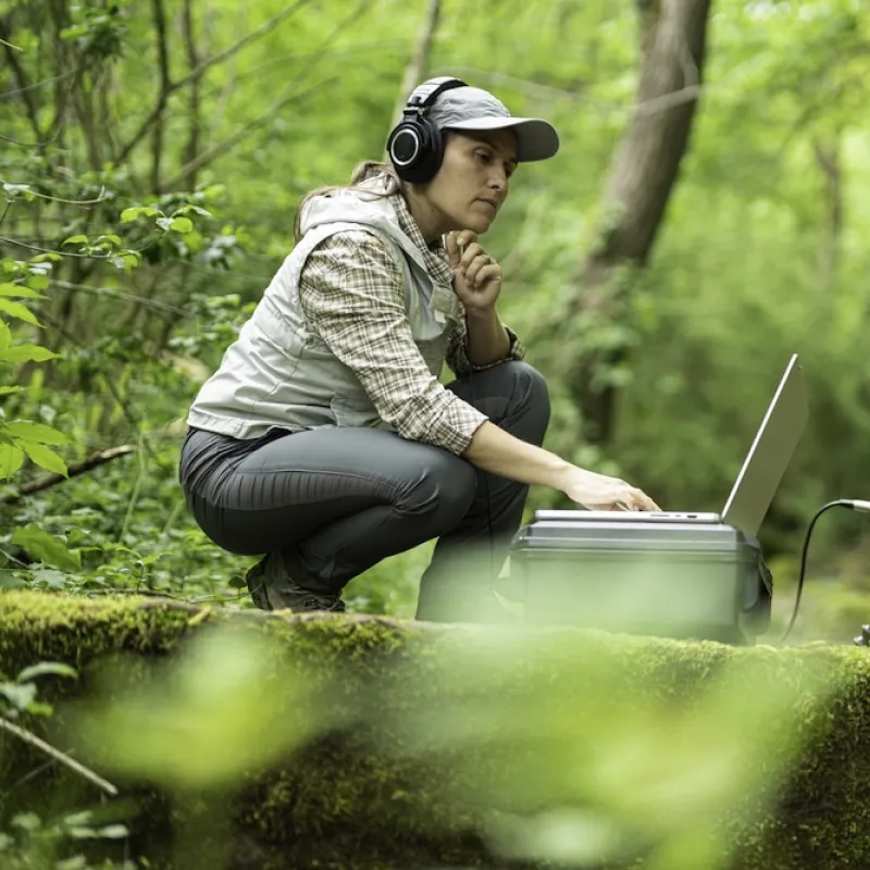 Female presenting person in bright green woods with audio recording equipment.