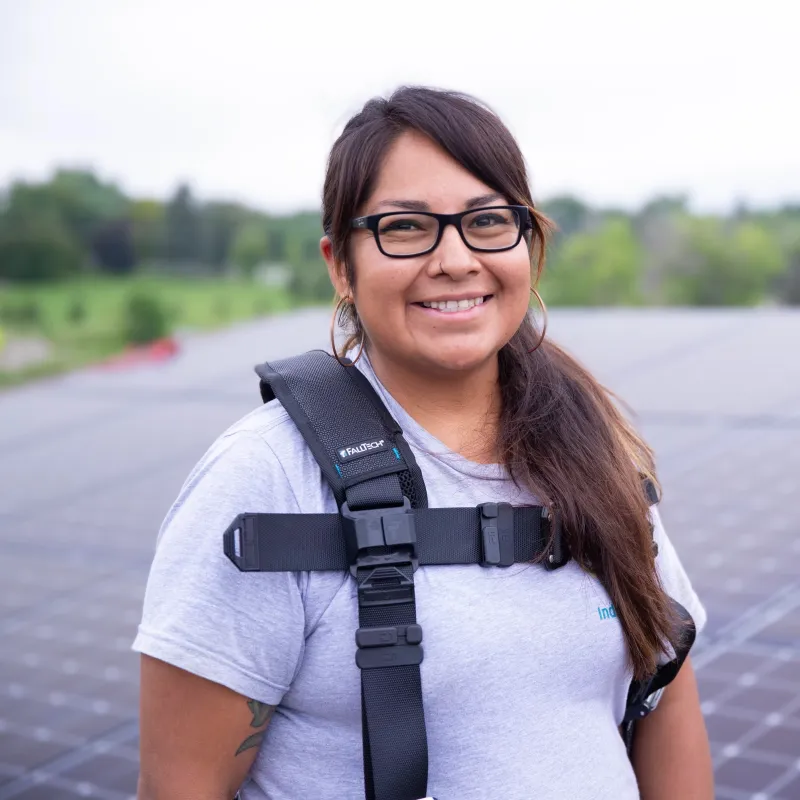 A portrait of a smiling, female presenting person with long brown hair and brown skin, wearing a safety harness, standing on a roof in front of installed solar panels.