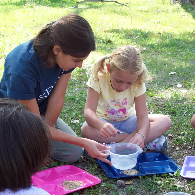 An environmental educator helps a student explore bugs while sitting in the grass under a tree